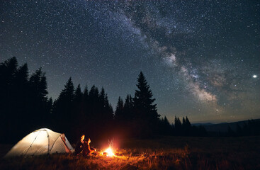 Side view of loving couple sitting near bright burning campfire and tent, enjoying beautiful camping night together under dark sky full of shiny stars and bright Milky Way, warm summer night. - obrazy, fototapety, plakaty