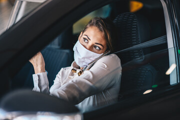 Young attractive fashionable businesswoman with protective face mask on driving herself on work while talking on a phone during corona virus outbreak.