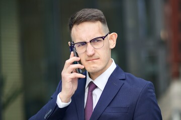 Portrait of angry annoyed young man talking on cell mobile phone. Upset businessman in formal suit and glasses having negative conversation on his smartphone, wrinkles his face