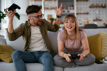 Boyfriend and girlfriend playing video game with joysticks in living room. Loving couple are...