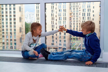 Girl gives five to boy. children sit on floor by window in new house during quarantine. Photo of children leisure house. Brother and sister are having fun at window. High five! 