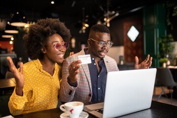 Attractive smiling african couple sitting in cafe and purchasing over internet things for house.