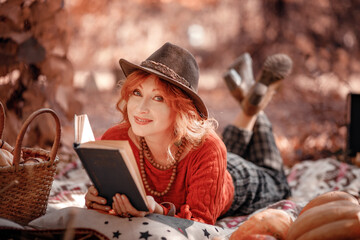 Fototapeta premium Autumn portrait of a red-haired woman in hat lying on a plaid in the park