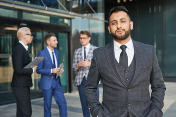 Portrait of young leader in elegant suit looking at camera while standing outdoors with his team in the background