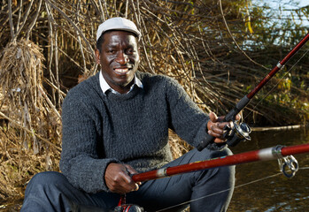 Fototapeta premium Portrait of cheerful afro fisherman sitting on wooden stool and fishing
