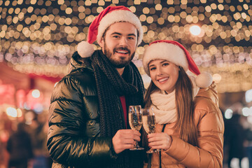 Photo of two people couple toast champagne glass 2021 x-mas greeting under evening outside illumination wear santa claus cap