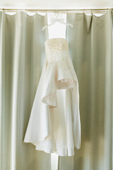 White dress of the bride of the original design on a hanger with a bow tie of the groom against the background of curtains.