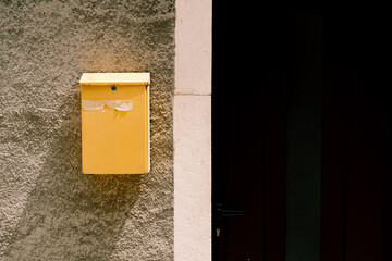 A yellow mailbox on a sunny day on a stone wall near the door to the house.