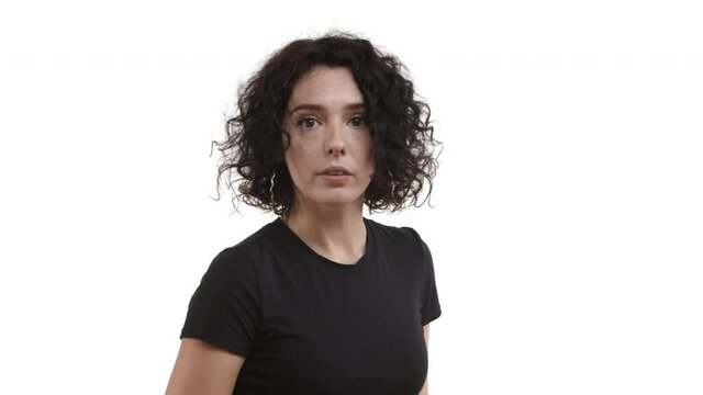 Annoyed and bothered woman with curly hairstyle, wearing black t-shirt, shushing at camera with finger pressed to lips, tell to keep quiet, standing over white background