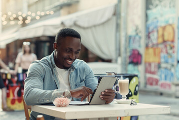 Young happy african american man uses tablet pc sitting on street cafe with graffiti on backdrop.