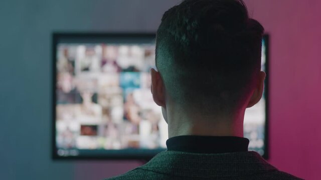 Rear view of man surfing watching pornography site on the web looking for virtual sex at night in a dark room in neon lights. Concept of porn addiction