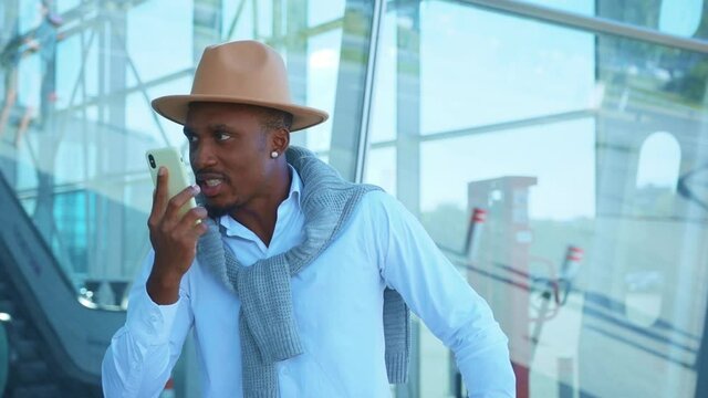 Face angry handsome African American in hat stand speak on phone near Airport feel nervous. Vacation and travel. Conversation outdoors stylish trip. Close up. Slow motion