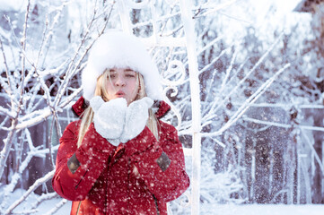 A girl in winter clothes blows snow off her mittens. portrait of a blonde in a winter fur hat.