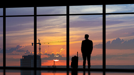 Fototapeta na wymiar The man with a suitcase standing near a panoramic window against the city sunset