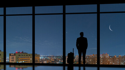 Fototapeta na wymiar The man with a suitcase standing near a panoramic window against a night city