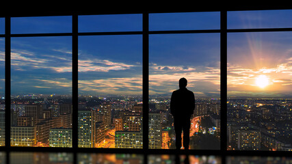 The male standing near the panoramic window against the bright sunset - obrazy, fototapety, plakaty