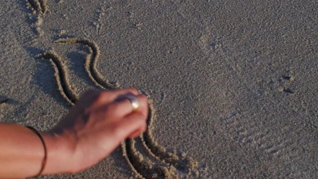 Slowmotion of young woman writing the word of Love on the sand sea beach. Girl draw on the sandy near waves in warm sunlight of early morning. Summer vacation concept.