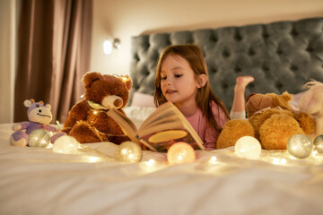 A cute girl is lying on her stomack in a huge bed barefoot reading a book having her teddybears and...