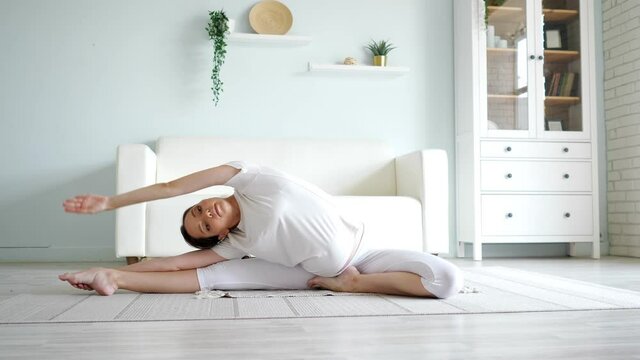 Attractive pregnant lady in white clothes does janu sirsasana position sitting on floor with rug against designer couch in spacious room at home