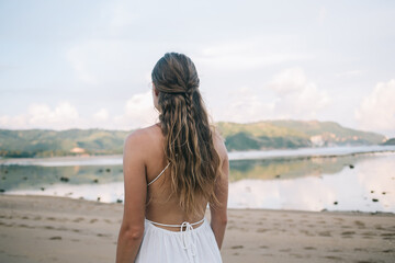 Peaceful braided woman in white dress walking along beach