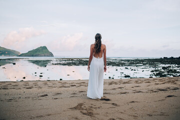 Tanned braided woman in white dress walking along beach