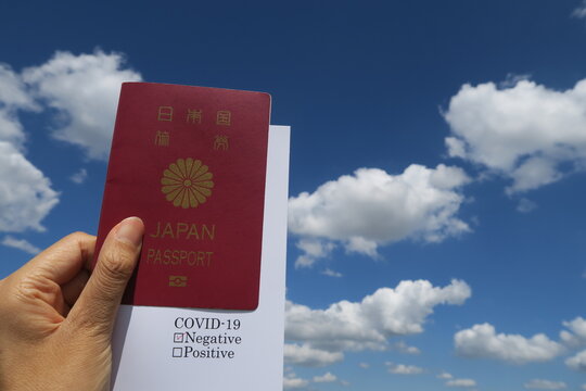 日本国のパスポートとコロナウイルス陰性にチェックの入った白いカードを持つ女性の左手。背景は青空と白い雲。