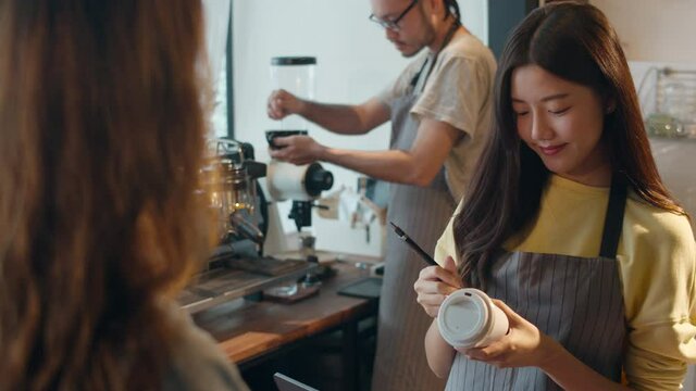 Young Asia female barista waiter taking order from customer standing behind bar counter while talking with customer making note on takeaway coffee cup at cafe restaurant. Owner small business concept.