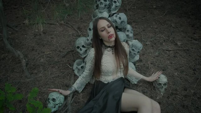 portrait of a young woman vampire, beautiful red lips with dripping blood drops on her face, sharp false fangs. A model in a dark gothic forest against the backdrop of the scenery of human skulls.