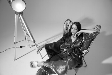 Young fashion female model with long straight brunette haie and makeup with pink eye shadows wearing silver kimono with trousers and white clog shoes sitting with crossed legs in stylish armchair