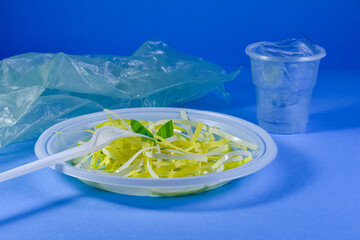Disposable plastic tableware, plate, fork, knife, glass on a blue background. There is plastic food in the plate. Composition for environmentalists. Healthy eating.