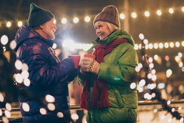 Photo of two people pensioner couple hold mugs drink eggnog comfortable cheerful smile newyear atmosphere wear mittens coat red scarf headwear x-mas night street park lights fair outside