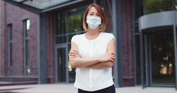 Confident businesswoman in medical mask standing in office buildings district while pandemic period. Good-looking Caucasian female worker crossing arm and looking at camera. Coronavirus concept.