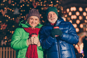 Two old people woman man have x-mas christmas jolly time night walk hold hot warm takeout beverage mug in city center outside under illumination wear season outerwear scarf hat