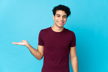 Young Venezuelan man isolated on blue background holding copyspace imaginary on the palm to insert...