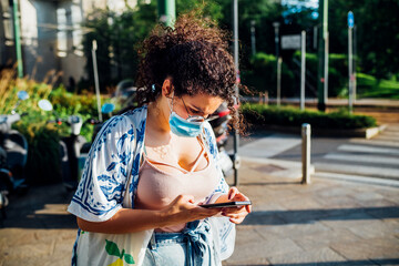 Multiethnic young woman wearing mask using smartphone outdoor - Mixed race young adult female in the city holding smartphone surfing internet