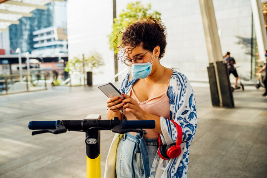 Young Multiethnic Woman Wearing Mask Unlocking Escooter - Diverse Ecological Female Outdoor Using Smartphone Driving Kickscooter