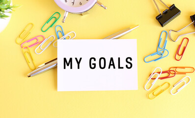 Top view of a business card with text MY GOALS, pen, paper clips and clock on a yellow background. Business concept.