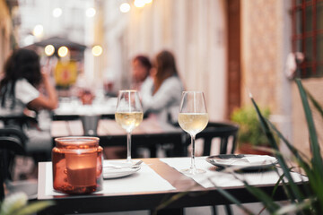 Two glasses of white wine, candles on a served table in an outdoor terrace, romantic dinner date with evening cozy lights