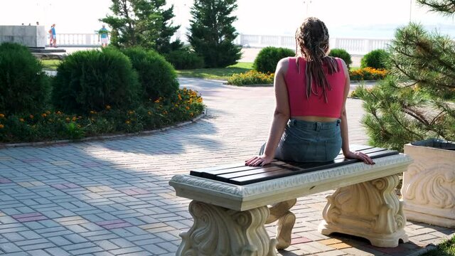 Young girl enoying summer green bushes and the sea. Concept. Rear view of a woman with many braids sitting on a white marble bench in front of a beautiful embankment.