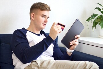 Young handsome man sitting on sofa at home in living room with tablet and credit bank card. Online payment in internet shop concept