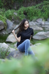 The blonde girl sits on a rock wearing a black shirt while stylish with views of natural rocks, lake water and clear sky. female t-shirt models for mockups and templates.