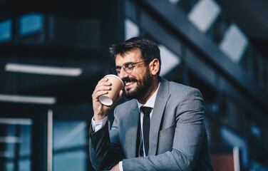 A young businessman stands in front of the company, drinks coffee and smiles