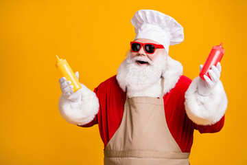 Fototapeta na wymiar Photo of positive santa claus chef hold mustard tomato ketchup sauce bottle x-mas eve noel snack wear headwear sunglass apron isolated over yellow shine color background