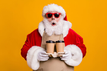Close-up portrait of his he nice attractive amazed stunned Santa waiter wearing apron holding in hands two cups order latte cappuccino isolated bright vivid shine vibrant yellow color background