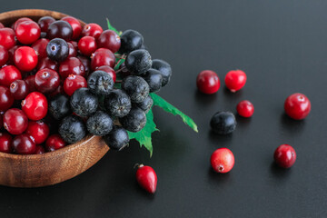 Cranberries and chokeberry in wooden bowl on black background. Nature, autumn, crop, food, berry concept.