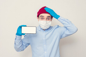 Shocked man in a medical mask shows the phone, grabs his head, portrait, copy space