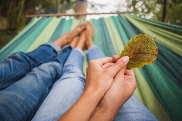 Obraz premium man and woman in love lying in hammock embracing, legs, close-up details, romance, autumn vacation, forest, barefoot, holding leaf in hands together, jeans, denim outfit