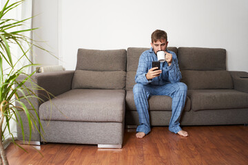 Obraz premium Young man dressed in pajamas at home having coffee while checking the smartphone.