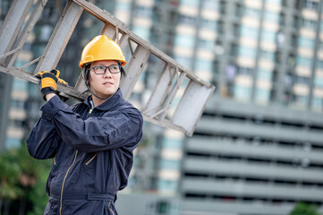 Asian maintenance worker man with protective suit and safety helmet carrying aluminium step ladder at construction site. Civil engineering, Architecture builder and building service concepts