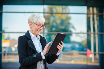 Smiling and happy senior businesswoman using digital tablet on the street in front of an office building